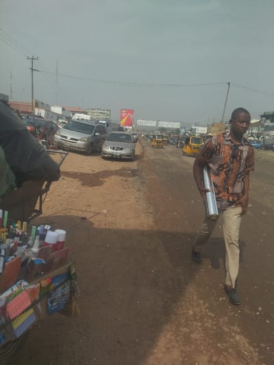 The image depicts a street scene in Way, Nigeria, taken at the coordinates latitude: 9.029, longitude: 7.584. A man is walking down a dirt road, carrying a large, silver, cylindrical object. He is wearing a brown shirt with a patterned design and beige pants. To the left of the road, there are several parked cars, including a silver minivan and a silver sedan. There is a vendor's cart parked on the side of the road, filled with various items. There are several other vehicles in the background, including motorcycles, cars, and trucks. There are power lines running overhead. The road is dusty and appears to be unpaved. The sky is clear and hazy, with very little cloud coverage. The overall impression is of a busy, bustling urban scene.