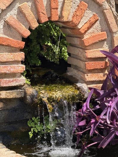 This close-up shot captures a decorative garden feature, likely a small water fountain or a cascading plant display. The primary subjects are the natural elements of water and plants, contrasted with man-made brickwork. A segment of a fountain's stone structure is visible, with water actively flowing down its surface, creating a shimmering effect and small bubbles. Growing from the dark, damp stone and near the water's edge are clusters of vibrant green, delicate ferns. Above the water and stone, bright purple leaves of a Tradescantia zebrina (also known as wandering dude or inch plant) cascade downwards, adding a striking color contrast.

The brickwork forms a semi-circular or spiral pattern, with rough, textured bricks in varying shades of red, orange, and yellow. These bricks are set against a lighter, granular mortar or concrete, suggesting a deliberately constructed, artistic element within the garden. The sunlight suggests it is daytime, likely outdoors in a garden or patio setting. The overall mood is tranquil and natural, highlighting the interplay of water, greenery, and rustic textures. There are no visible people or text in the image. The location context of Benalmádena, Spain, suggests a warm climate where such lush greenery and outdoor water features are common.