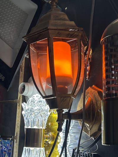 The scene depicts an assortment of illuminated and unlit lighting fixtures on display in Алат, Uzbekistan. Central to the image is an ornate wall-mounted lantern-style light fixture, finished in distressed bronze or gold, featuring clear glass panels that enclose an internal cylindrical LED bulb emitting a warm, flickering orange light, simulating a flame. To the upper left, a portion of a rectangular white LED panel light is visible, displaying numerous individual light-emitting diodes. Below this, a white ceramic Edison screw-type light socket is observed without an inserted bulb. In the lower left quadrant, a decorative clear glass or crystal light component with a fluted design and a metallic band is present. To the right of the central lantern, a metallic, vertically oriented light fixture, possibly a bug zapper or a decorative LED tube, emits an orange glow through a textured, grid-like surface. Numerous black electrical cables are visible, weaving between the various fixtures. The background is predominantly dark, suggesting an indoor retail or showroom environment for lighting products.