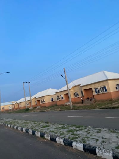 This media file captures an outdoor scene in Dumnga, Nigeria, likely during the late afternoon or early evening due to the soft, diffused light and clear blue sky. The perspective is a low-angle shot looking upwards and to the right.
The primary subjects are several multi-story residential buildings with light yellow and brown facades, featuring multiple windows. In front of one of the buildings, on a paved walkway, are a few individuals. Two of them are standing, and one appears to be sitting or crouching. They are small figures within the frame, suggesting a sense of scale.
Adjacent to the buildings is a paved road with white lane markings. A sidewalk with a decorative pattern of interlocking gray pavers and patches of green grass runs along the edge of the road. A distinctive curb with alternating black and white stripes borders the sidewalk. Overhead, a series of electrical wires stretch across the sky, supported by utility poles. One pole with a street lamp is visible.
The scene appears to be a residential area, possibly a housing estate. The weather is clear and pleasant, with no visible clouds. The overall mood is tranquil and ordinary, depicting a typical day in the community. There is no visible text in the image.