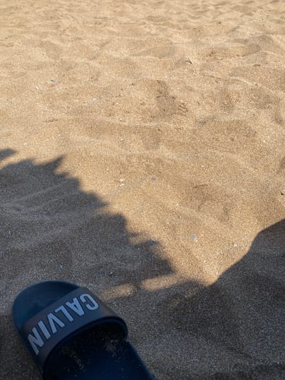 The image shows a close-up view of sandy beach in Panormos, Greece. A black sandal with "CALVIN" written on it in grey letters is visible in the bottom left corner of the frame. Shadows of palm trees are cast across the sand, suggesting a sunny day. The sand is light brown and has a granular texture with small pebbles and shells mixed in. The overall scene conveys a relaxed, tropical atmosphere.