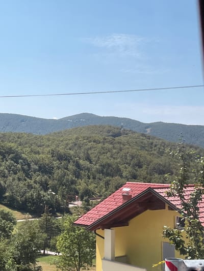 The image depicts a natural landscape with a bright blue sky and distant, verdant hills. A yellow house with a red tiled roof is prominently featured in the foreground, partially obscuring the view of the hills behind it. The house has a chimney and a white pillar. Some trees are visible in front of the house and scattered across the landscape. There are also power lines running horizontally across the upper portion of the image. The scene is likely located in or around Col, Slovenia, given the context of the request.