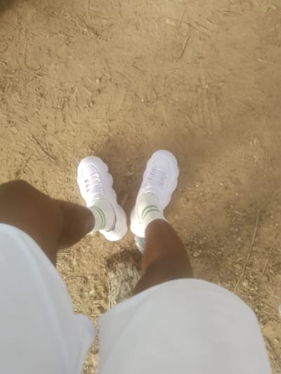 The image shows a close-up, overhead view of a person's legs and feet wearing white sneakers with green stripes on the socks. The person is standing on a dry, sandy ground with some scattered dried grass and small stones. The ground appears to be outdoors, possibly a path or an open area. The lighting suggests it is daytime. The person is wearing white clothing on their legs, which could be shorts or pants. There are some faint shoe prints visible in the sand. The location is stated to be in Majemu, Nigeria.