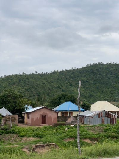 This media file shows a collection of houses with corrugated tin roofs and some with blue and white roofing, situated on a grassy hillside. A dense, green, tree-covered mountain rises in the background. The sky is overcast with a few wispy clouds. Two goats are visible in the foreground, grazing on the vegetation. A rustic wooden pole stretches horizontally across the scene, with wires attached to it. The houses appear to be simple structures, possibly in a rural or semi-rural setting in Kona Garu, Nigeria. The overall impression is one of a quiet, overcast day in a natural landscape with human habitation. There is no visible text in the image.