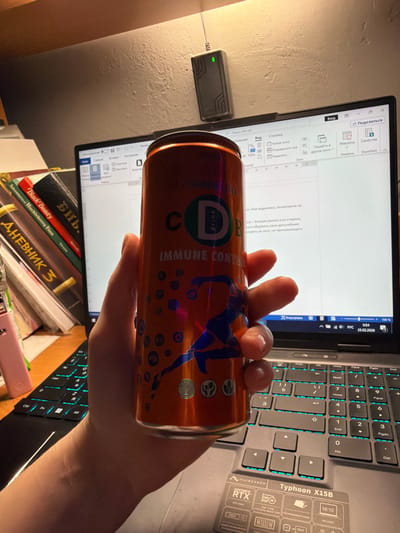 This forensic image captures an indoor desk scene, located in Lubnya, Russia. The primary subject is a human hand holding an orange beverage can. The can is metallic orange, featuring a prominent blue silhouette of a running figure, numerous small blue circles, and text including "C D drink" and "IMMUNE CONTROL." The hand grasps the can, with visible reflections on the fingers and fingernails.
In the background, a grey MAIBENBEN Typhoon X15B laptop, specified with an NVIDIA GeForce RTX 4060, is open. Its keyboard exhibits cyan-blue backlighting. The laptop screen displays a document, likely in Microsoft Word, with visible Russian text. The Windows taskbar in the lower right corner shows the date "25.02.2026" and the time "0:54."
To the left of the laptop, a stack of books is present, including titles in Russian such as "ДНЕВНИК" (Diary) and "Библ." (likely Bible), alongside English titles like "Black Beauty" and "Huckleberry Finn." A pink, slender object is partially visible near the books.
Mounted on the light-colored wall above the laptop is a black, rectangular electronic device with a single illuminated green LED, suggesting active operation, possibly a network repeater or similar peripheral. The area is illuminated by an unseen warm light source, casting a glow on the wall and desk.