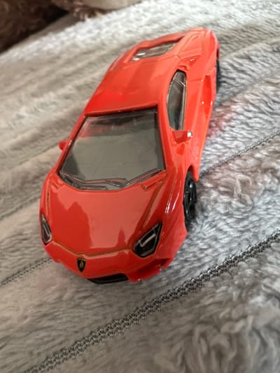 The image displays a red toy sports car, resembling a Lamborghini, positioned diagonally on a grey, textured, fibrous surface. The front of the vehicle points towards the lower-left quadrant of the frame. The toy features a red body, black wheels, transparent windows, and dual windshield wipers molded into the front windscreen. A Lamborghini emblem is visible on the front grille. The grey surface, likely a textile, exhibits faint, parallel patterns of linear stitching. A blurred, brown object occupies the upper-left background. No individuals are present, and no actions or interactions are depicted. The precise location within Easingwold, United Kingdom, is not discernible from the image content.