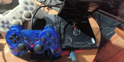 The image shows a close-up, overhead view of a PlayStation 2 console with its lid open, revealing the disc drive mechanism. A transparent blue PlayStation controller is positioned in the foreground, partially obscuring the console. To the left and slightly behind the blue controller is a white PlayStation controller, appearing older. The objects are placed on a wooden surface, possibly a table. The overall scene is dimly lit, with some dust visible on the console and controllers, suggesting the items may not be in frequent use. The location is not discernible from the image itself, and therefore the city of Есипово, Russia, cannot be determined from visual cues alone.