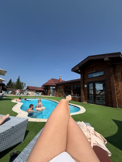 The image shows a sunny outdoor scene at a resort with a swimming pool. In the foreground, a person is relaxing on a lounge chair, with their legs prominently featured. In the middle ground, a kidney-shaped swimming pool is filled with bright blue water. Several people are in the pool: two men and a woman with a child. One of the men is talking to another man, while the woman and child are near an inflatable ring. In the background, there are wooden buildings with red roofs, an artificial turf lawn, and some trees. The sky is a clear blue. The location is identified as Staroabashevo, Russia, though this cannot be visually confirmed from the image itself.
