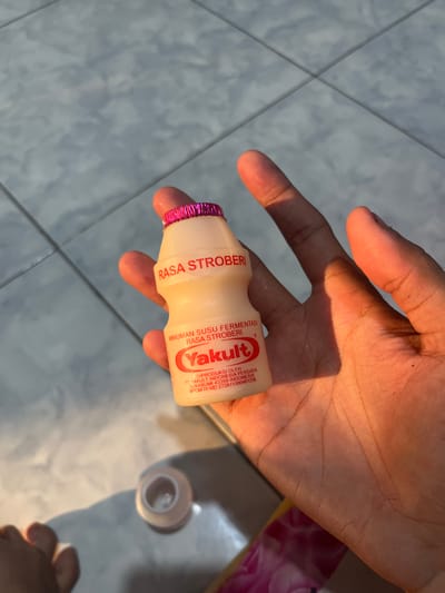 The image shows a person's hand holding a small, pink-capped bottle of Yakult fermented milk drink. The hand is illuminated by bright, natural light, casting distinct shadows onto a light grey tiled floor. The tiles form a square pattern with visible grout lines. In the lower right corner, a white plastic cap or lid is partially visible on the floor, slightly out of focus.

The bottle's label is clearly visible, identifying the product as "MINUMAN SUSU FERMENTASI" (Fermented Milk Drink) and specifying "RASA STROBERI" (Strawberry flavor), which is also indicated by the pink cap. The brand name "Yakult" is prominent. Further text details the producer: "DIPRODUKSI OLEH: PT YAKULT INDONESIA PERSADA SUKABUMI 43359 INDONESIA" along with a BPOM RI registration number "MD 272811000401236". The bright lighting suggests it is daytime. The scene appears to be indoors, likely in Paweden, Indonesia, as per the provided location context.