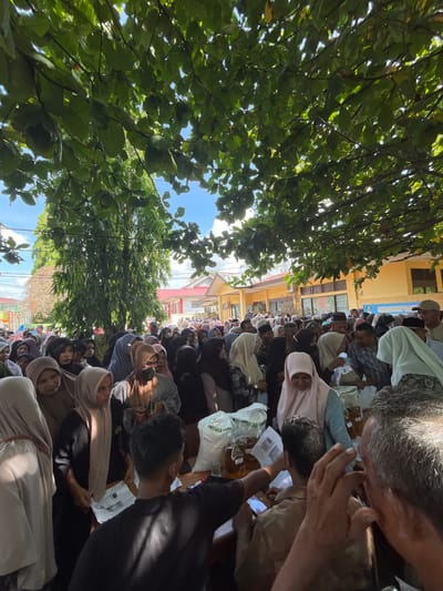 In Pulodolang, Indonesia, a crowd of people are gathered outdoors under the shade of large green trees. The crowd appears to be participating in some form of distribution or event, as many individuals are holding bags and paper documents. Several large, clear plastic bags filled with a yellowish liquid are visible on tables, along with sacks of what looks like rice. The people are dressed in a mix of casual clothing and headscarves, suggesting a diverse group. In the background, a yellow building with red trim is visible, possibly a community center or administrative building. The scene is illuminated by bright sunlight, indicating a daytime event. The overall impression is one of a community gathering for a distribution of goods.