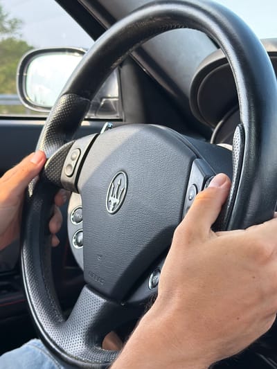 The image shows a close-up of a person's hands on a black steering wheel inside a car. The steering wheel has perforations and a silver Maserati emblem in the center. There are buttons on the steering wheel, some of which are labeled "INFO". The side mirror of the car is visible in the upper left corner, reflecting a blurry outdoor scene with trees and sky. The image is taken from a driver's perspective. The location is not determinable from the image content.
