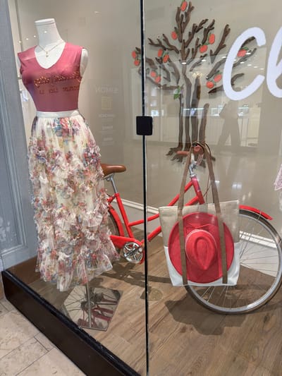 In a storefront window display in Mission Viejo, California, a female mannequin is adorned in a pink, sheer top with gold lettering and a long, flowing white skirt featuring a floral print and ruffled tiers. To the right, a bright red bicycle is parked, with a canvas tote bag carrying a red hat resting on its rear. The background of the display includes a decorative tree with red embellishments resembling fruit and a reflection of the store's interior, showing shelving and a person walking.