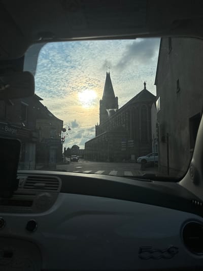 The image, captured from inside a vehicle, depicts a street scene in Saint-Maclou-de-Folleville, France, likely during the daytime with the sun setting or rising. In the foreground, the dashboard of a white Fiat 500 is visible, including the steering wheel, dashboard vents, and a silver "500" emblem.
The view through the windshield shows a street lined with buildings. On the left, a building with a sign reading "- Burger" is partially visible. Next to it, another building has a sign for "LA CIVETTE." Further down the street, a large, dark-colored church with a tall spire dominates the skyline. The church exhibits Gothic architectural features, including pointed arches and stained-glass windows. A statue of a figure holding a cross is visible atop one of the church's roofs.
Several cars are present on the street, including a white sedan parked near the church and a dark SUV further in the distance. The sky is filled with scattered, puffy clouds, with the sun creating a bright halo effect. Street signs and traffic lights are also discernible. The overall impression is a quiet, somewhat overcast day in a French town, with the church serving as a prominent landmark.
