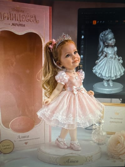 The image displays a doll dressed as a princess, positioned in the foreground. The doll is wearing a tiara and a light pink, frilly dress adorned with a bow. The doll stands on a white circular platform. To the left of the doll is a pink box with Russian text that translates to "Fashionable Princess" and "Dreams." Further left, a circular emblem reads "Made with love." Behind the doll, to the right, is a gray figurine of a similar doll, and a decorative glass bottle. A light pink rose is visible in the bottom right corner. The background appears to be a studio or display setting. The image does not provide any information or visual cues that would allow for its location to be identified as Protasovo, Russia.
