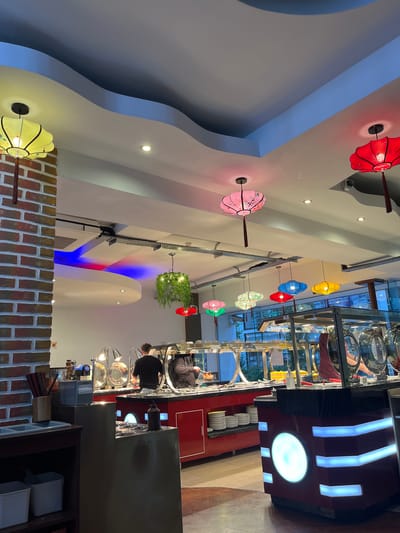 The image depicts the interior of a restaurant, likely in Eastbourne, United Kingdom, judging from the architectural style. The focus is on the buffet area, where patrons appear to be serving themselves. Overhead, there is a decorative arrangement of colorful lanterns and a hanging plant. The ceiling has a unique, layered design with blue accents. The restaurant is equipped with modern serving stations, a display of plates, and a well-lit environment. The foreground includes a brick wall detail.