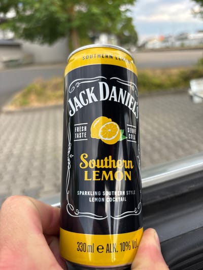 This media file is a close-up, vertical shot of a can of Jack Daniel's Southern Lemonade, held by a person's hand. The can is black with yellow accents and features the Jack Daniel's logo, an illustration of lemons, and text indicating "Southern Lemon," "Sparkling Southern Style Lemon Cocktail," "Serve Cold," "Fresh Taste," and "330 ml e ALK. 10% Vol." The background is blurred, showing hints of an outdoor setting with greenery, paving stones, and what appears to be a road or pathway, suggesting an outdoor location. The lighting suggests it is daytime. The person's hand is visible on the right side of the frame, holding the can. The overall impression is of a casual, outdoor moment with a beverage.