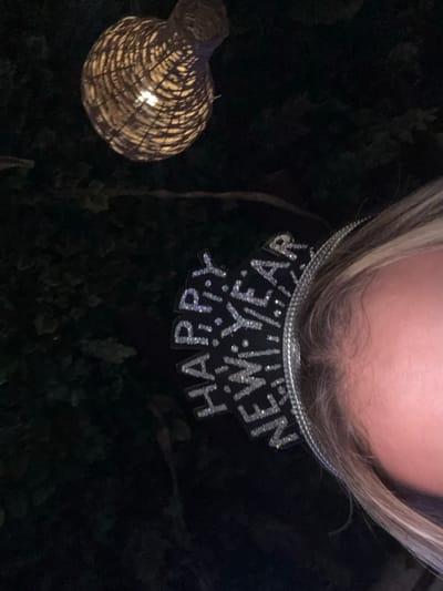 A person wearing a "Happy New Year" headpiece is angled downward. Only the top of their head and the headpiece are visible. To the left, there's a woven lantern emitting a soft glow. The background is a mix of darkness and foliage. The image is taken at an unusual angle, looking down and slightly off-center.
