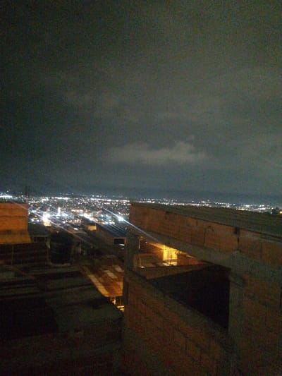 A nighttime view overlooking a sprawling city with distant lights stretching to the horizon beneath a cloudy sky. In the foreground, weathered brick structures and rooftops suggest an elevated vantage point, possibly a building under construction or a similar urban element. The scene is captured in low light, with details obscured by shadows, enhancing the mystery and urban atmosphere.