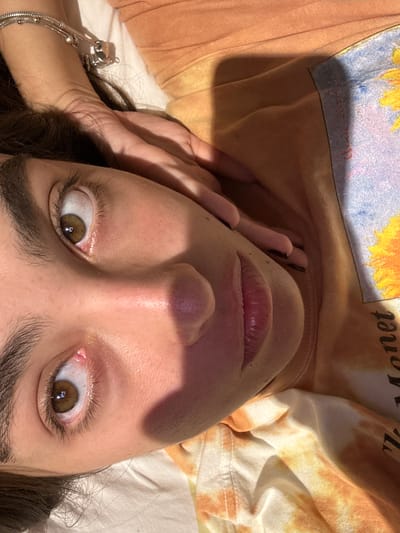 A close-up, tilted view of a person's face looking upwards. The person has fair skin, brown eyes, dark eyebrows, and slightly parted lips. A hand with painted black nails rests near the left side of the face. The person is wearing a t-shirt with a tie-dye pattern in shades of orange, yellow, and white. The image is lit by sunlight, creating shadows across the face. A silver bracelet is visible on the person's left wrist. The location is unknown and not discernable from this image.
