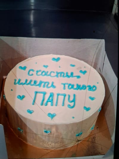 The image shows a round cake decorated with blue frosting. The cake is placed on a gold circular board. The cake has a light pink frosting and is adorned with blue icing details in the shape of hearts and dots. Text in blue icing is written on the cake in Russian. The text reads "Счастья иметь такого ПАПУ" which translates to "Happiness to have such a DAD". The scene is indoors, with the cake on a table. The lighting is artificial, suggesting an indoor event or preparation. The focus is on the cake itself, highlighting the decorative elements and the inscribed message.