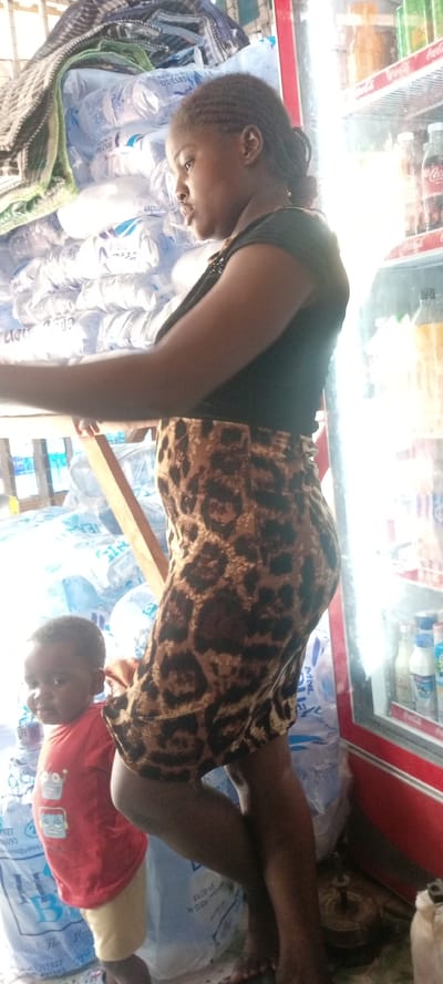 The image captures a scene inside a small shop or stall, possibly in Kwachiri, Nigeria, featuring an adult woman and a young child.
The woman is positioned towards the center-right, seen in profile, facing left. She has dark skin and braided hair styled back. She wears a black short-sleeved top and a knee-length, form-fitting skirt with a brown and black leopard print. Her arms are slightly extended forward, suggesting she is reaching or performing a task. Her facial expression appears neutral and focused.
To her left, large clear plastic bags filled with bottled water are stacked high, creating a backdrop. Visible text on these water bags includes partial numbers and letters such as "D1-1585", "D.0115", "REG N", and "The B". A patterned blanket or cloth is draped over the top of the water bottle stacks. A wooden beam or structure is also visible behind her.
To the woman's right, a red refrigerator with glass doors stands, brightly lit from within. It is stocked with various bottled beverages, some of which bear "Coca Cola" branding.
At the bottom left, a small child, likely a toddler, is partially visible, looking towards the viewer. The child is wearing a red t-shirt with a robot design and light-colored shorts, standing near the stacked water bottles and the woman's leg.
The bright lighting suggests it is daytime. The setting appears to be an active, functional retail space, possibly a convenience store or a vendor's stall.