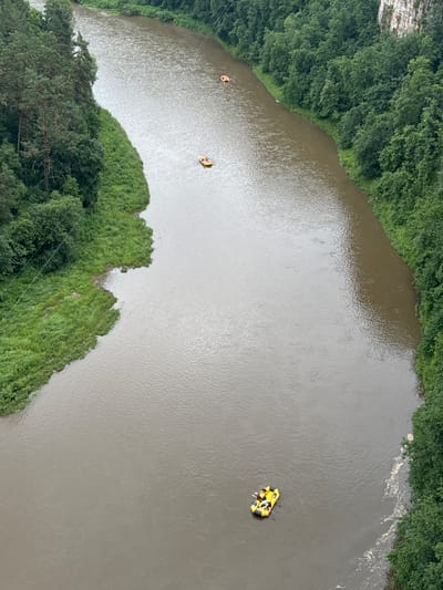 The image shows an aerial view of a wide, murky river bordered by lush green trees and vegetation. The river is not perfectly still; there are ripples and some swirling patterns on its surface, suggesting a gentle current. Along the banks, dense green foliage covers the land, with some areas of lighter green grass leading down to the water's edge.
In the river, there are two inflatable rafts with people on them. One raft, closer to the left bank, is yellow and orange and appears to have at least one person in it, who is wearing an orange life jacket. Further out in the river, a second, larger yellow raft is visible with multiple people inside. They are paddling, indicating that they are engaged in a recreational activity like rafting or kayaking.
The overall impression is of an outdoor recreational scene in a natural environment. The lighting suggests it is daytime, possibly overcast given the muted light and lack of strong shadows. The dense greenery implies it is summer or late spring. There is no visible text in the image. The location context provided is Kul'metovo, Russia.