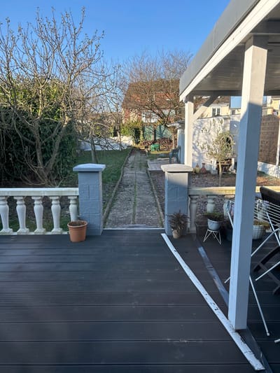 The image presents a residential backyard in Saint-Quentin, France, viewed from a dark grey composite deck on a clear, sunny day. The foreground consists of horizontal composite decking planks. To the left, a white balustrade with several balusters is adjacent to a grey block-textured pillar. An empty terracotta pot sits on the deck near this pillar. A corresponding grey pillar is located to the right, framing the entrance to a garden path. Next to the right pillar, a potted plant with dark, possibly fern-like leaves is visible. Two long, narrow strips, one white and one dark, lie on the deck to the right, extending towards a white structural post supporting a light-colored pergola roof. Partially visible to the far right are a white metal clothes drying rack and a brown folding chair.
The midground features a narrow, unpaved path with concrete sections and interspersed grass, extending into the background. To the left of the path, dense, leafless trees and green foliage line a boundary. To the right, a white wall parallels the path, with a small tree (possibly an olive tree) in a pot and several other decorative pots placed against it. A small wooden platform is visible further along the path on the right.
The background shows a garden area sloping slightly upwards. More bare trees are present, and a green plastic chair is visible in the distance on the right. Residential buildings with reddish-brown tiled roofs and light-colored facades are discernible beyond the garden, under a cloudless blue sky. No individuals or active subjects are present in the scene.