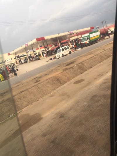 A gas station is visible alongside a roadway. It has multiple pumps under a wide canopy. Several vehicles are parked nearby, including a white van and a large green and white truck. Some people are standing near the vehicles and pumps. Electrical poles are seen in the background. The scene appears to be viewed from inside a car, with a portion of the window frame visible. The sky is overcast.