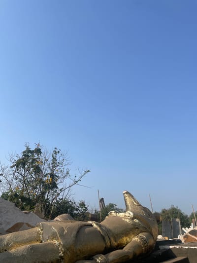 The image depicts a golden Anubis statue in Mira-Bhayander, India. The statue is positioned on a white base, surrounded by foliage. The backdrop is a clear, light-blue sky.