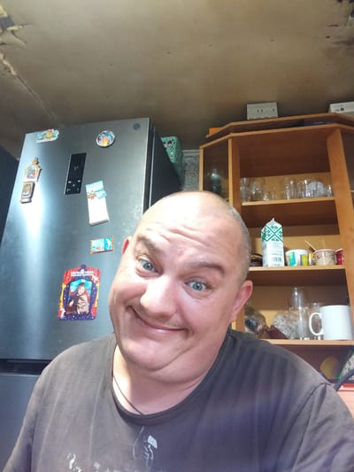 A man with a shaved head and blue eyes is taking a selfie in what appears to be a kitchen. He is wearing a dark t-shirt. Behind him, to the left, is a silver refrigerator with several magnets on it, including a framed photo of a boy with a dog. To the right is a wooden shelf unit filled with various items, including glasses, cups, and a carton of milk. The ceiling above appears to be textured and possibly damaged. The image is taken from a slightly low angle, looking up at the man. There is no indication of the city of Sloboda, Russia, or any other location.