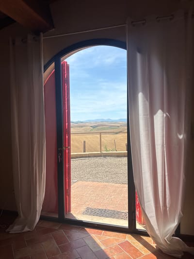 This is a horizontal, eye-level, medium shot of an open, arched doorway leading to a scenic outdoor view of rolling hills under a bright blue sky. The doorway is framed by red-painted double doors, one of which is ajar. White curtains hang on either side of the doorway, partially obscuring the view. The interior floor is tiled with terracotta-colored squares and rectangles, with sunlight casting diagonal shadows across it. Outside the doorway, a gravel pathway leads to a red-tiled patio. Beyond the patio, the landscape features dry, golden fields, some with visible rows, and a wooden fence. In the distance, rolling hills are visible, with a hazy mountain range on the horizon. The scene is devoid of people or animals. The overall impression is one of serene Tuscan countryside in Lajatico, Italy.