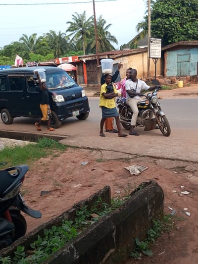 This image captures a vibrant roadside scene in Oko Town, Nigeria, illustrating local commerce, human activity, and urban infrastructure. The foreground features unpaved reddish-brown earth and a concrete drainage ditch with green vegetation. An asphalt road traverses the midground, bordered by a concrete curb. In the background are several single-story buildings, some with corrugated metal roofs and peeling paint, indicating both commercial and residential use. Lush green foliage and palm trees punctuate the skyline. A utility pole stands on the right, next to a signpost displaying text including "INSTITUTION OF AGE." The sky is overcast.
Four individuals populate the midground. A young male, in a dark t-shirt, light brown patterned shorts, and flip-flops, stands next to a dark commercial van, balancing a clear plastic container with a white granular substance on his head. A young female, wearing a yellow short-sleeved top, blue skirt, and flip-flops, also carries a similar container on her head and holds a black plastic bag. An adult male, in a white polo shirt, light khaki trousers, and dark flip-flops, is seated on a light-colored chrome motorcycle, carrying a dark bag across his chest, looking left with a slight smile. Partially obscured behind him, an adult female in a pink garment also looks in the same direction.
Key objects include a dark-colored commercial van (LND 111FS) with a partially visible red and white roof sign (possibly "TAXI") and a small white dashboard sign. The chrome motorcycle carries the seated male, while a partial frame and front wheel of another motorcycle are visible in the foreground. The two clear plastic containers on the individuals' heads hold a white granular substance. A yellow jerrycan is near a light blue background building. Other visible text includes "MIRA HOTEL" on a background structure. Scattered paper, plastic, and crumpled cardboard litter the unpaved ground. The younger individuals appear to be street vendors, and the group on the motorcycle seems to be engaged in an interaction or observing something to their left, typifying a lively moment of street life and commerce.