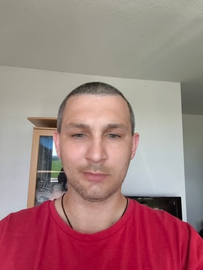 This is a close-up selfie of a man with a shaved head and light stubble. He is wearing a red t-shirt and a dark necklace. His eyes are light green. The background appears to be indoors, with a wall and a piece of furniture to his left, possibly a cabinet with glass doors. Behind him, to his right, is a television. The lighting suggests it is daytime. The setting is likely a home in Wolgast, Germany. No specific activity is apparent; the man is looking directly at the camera.
