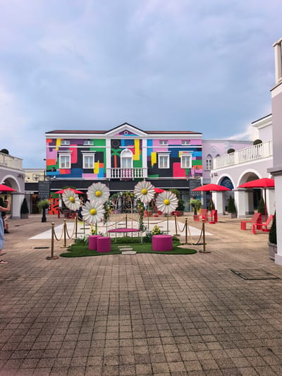 This media file depicts an outdoor courtyard area, likely a shopping outlet or entertainment district, in what appears to be a European setting, possibly Italy. The scene is dominated by a building with a striking, colorful facade adorned with geometric shapes in vibrant hues of pink, yellow, blue, green, and purple. This building features multiple windows and an ornate white railing on a balcony.
In the foreground, a decorative display is set up on a patch of artificial grass. This display includes large, white daisy-like flowers with yellow centers, a small ornate white gazebo covered in greenery, and two bright pink ottomans. Gold stanchions with rope mark off the decorative area. The ground is paved with textured square tiles.
Several red umbrellas and chairs are visible in the background, suggesting a seating area or outdoor cafe. To the left, a woman in a blue and white striped dress is partially visible, holding up a phone, seemingly taking a photo of the building. Other shoppers or visitors are also seen in the distance, browsing shops or walking through the courtyard.
The time of day appears to be daytime, with overcast skies indicating a cloudy or possibly impending rainy weather. The overall atmosphere is bright and cheerful, largely due to the colorful architecture and playful decorations, despite the subdued sky. The location context provided mentions "Aiello del Friuli / Dael, Italy," which, if accurate, places this scene in northeastern Italy.
A sign is visible on the building to the left of the colorful structure, displaying text that includes "MOSCHINO" in large lettering. Other signs are present but illegible due to distance and resolution.