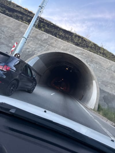 The image shows the entrance to a tunnel on a sunny day. A black SUV is parked to the left of the tunnel entrance, facing towards it. The tunnel is arched and made of concrete. There is a metal pole with a ladder on the left side of the tunnel entrance, and a red and white striped sign attached to it. The road leading into the tunnel is asphalt and has white lane markings. The surrounding area is hilly and covered with vegetation. The sky is blue with some clouds. The image appears to be taken from inside another vehicle, as a portion of a white car's hood is visible in the foreground. No people are clearly visible in the image.