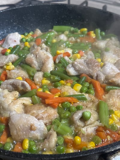 A dark-colored frying pan, positioned over a heat source, is observed in a close-up, overhead view within an indoor kitchen in Fowlis Easter, United Kingdom. The pan contains a mixture of diced white meat, consistent with chicken, intermingled with cut green beans, orange diced carrots, yellow corn kernels, and green peas. The ingredients are submerged in a visibly simmering, light-colored liquid, producing steam. The chicken pieces exhibit some surface browning. No individuals are present in the frame.