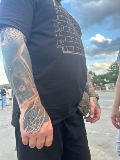 In Palanca, Moldova, a man with extensive tattoos on his arms and hand stands facing forward, partially obscuring a woman to his right. The man is wearing a black t-shirt with a blueprint-like design and black pants. He has a green fanny pack slung across his waist and a green watch on his left wrist. His left arm displays tattoos of a train and geometric patterns. His right hand, visible in the foreground, features a tattoo of a trident symbol. The background shows a paved area, trees, buildings, and a cloudy sky. Several other individuals are visible in the distance.