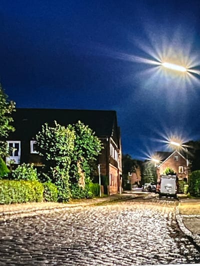 The image depicts a street scene at night in Nijmegen, Netherlands. The street is paved with cobblestones and lined with trees and hedges. On the left, a brick house with white-framed windows is visible. Further down the street, another house with a prominent gable and lit windows is seen. A white delivery van is parked on the right side of the street, facing away from the camera. The sky is dark blue, with bright light flares emanating from artificial light sources, suggesting streetlights or building lights. The overall atmosphere is serene and quiet, indicative of a residential area.