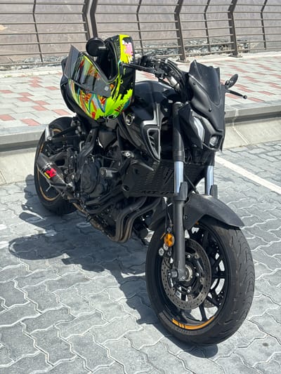 The image shows a black Yamaha MT-07 motorcycle parked on a paved surface. The motorcycle is oriented with the front wheel towards the upper right corner of the image, and the rear towards the lower left. A colorful helmet rests on the left side of the motorcycle. The background consists of a textured pavement and a metal railing. The location is Khor Fakkan, United Arab Emirates.