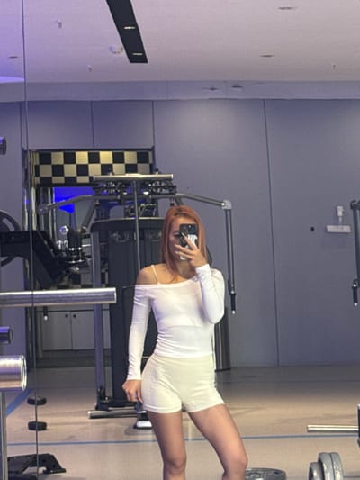 The image depicts a woman taking a mirror selfie inside a fitness facility located in Lishan District, China. The central subject is a woman, reflected from the waist up, holding a dark-colored smartphone that partially obscures her face. She has shoulder-length, reddish-brown hair. She is wearing a white, long-sleeved, off-the-shoulder top and light-colored athletic shorts. Her left shoulder is exposed, supported by a thin strap.

The background, visible around and behind the woman's reflection, consists of various gym equipment. On the left, several components of a cable machine are visible, including metal frames and weights. A black and white checkerboard pattern is present on an upper wall or surface section in the upper left quadrant. The walls of the facility are light gray or bluish-gray, with some panels. Overhead, the ceiling is light-colored, featuring recessed lighting and a dark, rectangular light fixture. In the lower right, several dark weight plates are stacked on the floor. The environment indicates a modern, well-lit indoor gym setting, characterized by reflective surfaces and exercise machinery. No other individuals are present in the observable scene.