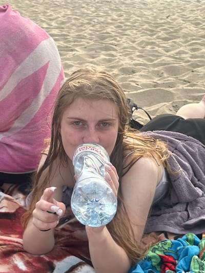 A young person with long, wet, blonde hair lies on a sandy beach, holding a plastic bottle of water to their lips. They are partially covered by towels, including a purple one draped over their back and a pink and white striped towel beneath their head. The sand is rippled, suggesting it might be windy or that the tide has recently receded. The person's eyes are open and they are looking at the camera. Their fingernails are painted white. The lighting suggests it is daytime, possibly late afternoon due to the warm tones. The overall mood is relaxed and casual.