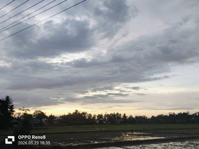 The image depicts a landscape in Kampungjumpa, Indonesia, at sunset. The sky is overcast with grey clouds, revealing a band of golden light along the horizon. In the foreground, a flooded rice field reflects the ambient light. Beyond the field, a dense row of trees and vegetation obscures the houses and structures. Power lines are visible in the upper left corner of the image. The photograph was taken on an OPPO Reno8, as indicated by the watermark, with the date and time stamp showing May 23, 2026, at 18:35. There are no people visible in the image.