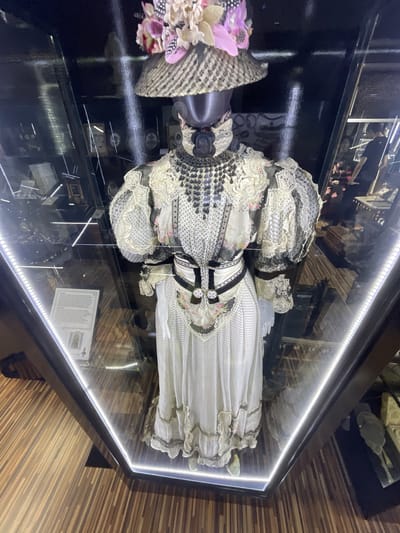 A vintage dress with ornate detailing is displayed in a glass case illuminated by LED lights. The dress is white with a pattern of small black dots and floral accents. It features a high neckline adorned with black beads and lace, puffed sleeves with lace trim, and a full skirt with ruffled hems. A wide black and white belt cinches the waist. A wide-brimmed hat, decorated with pink and white flowers and feathers, sits atop the mannequin. The display is situated on a wooden floor in what appears to be a museum or exhibition space. The city of Voskhod, Russia, is not visually depicted or mentioned in the image or its context.