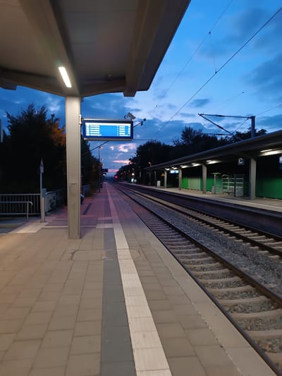 The image shows a train station platform in Germering, Germany, at dusk. The sky is a deep blue with some clouds, tinged with the colors of sunset. Overhead, power lines for the trains crisscross the sky. The platform is paved with square tiles and has a textured gray and white strip along the edge. A covered waiting area with lights runs along the platform on the right side, with green panels behind it. On the left side of the platform, a metal railing separates the platform from a darker, tree-lined area. A digital display board mounted on a pole shows train information. The train tracks run parallel to the platform, disappearing into the distance. The overall impression is one of quiet solitude, as the day ends and evening begins at a suburban train station.
