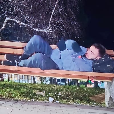 At night in an outdoor public area in Bachevo, Bulgaria, an adult male is observed lying horizontally on a multi-slatted wooden park bench. The individual, with dark hair and a beard, is dressed in a light gray tracksuit, black gloves, black socks, and black athletic shoes. He lies supine on the bench, head to the right, resting on a black and camouflage-patterned backpack utilized as a pillow. His eyes appear closed, indicating a state of rest or sleep. His left arm is bent with the gloved hand positioned near his chest. Bare tree branches are visible against the dark sky in the background. Below the bench, a grassy area is present with scattered white debris. A paved sidewalk is visible in the foreground to the left, and a piece of brown cardboard rests on the ground to the right.