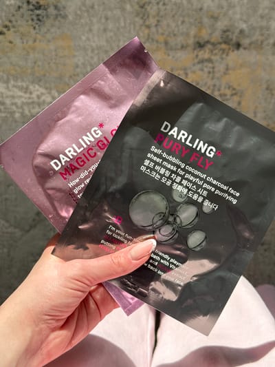 The image shows a close-up of a person's hand holding two Darling* brand sheet masks. The mask on top is black with silver text and images of bubbles, and it reads "DARLING* PURY FLY Self-bubbling coconut charcoal face sheet mask for playful pore purifying." The mask underneath is pink with pink and white text and reads "DARLING* MAGIC GI Glow rev-". Some of the English text on the black mask also says "I'm your fun-time friendly playmate for tickling far... bath with Vitamin E, and AGAVE extract putting P... w back into your face. ALLANTOIN Made in". The background appears to be a textured surface, possibly a rug or floor, with some bright light casting shadows. The lighting suggests it might be daytime. The scene is focused on the products and the hand.
