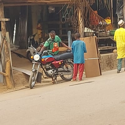 The image displays an outdoor street scene, likely taken during daytime. The perspective is slightly angled downwards, looking at people and objects situated on a street or path. The image is oriented such that the ground/road appears on the right side, extending vertically.
In the mid-ground, a person, possibly a young man or teenager, wearing a light green t-shirt with a prominent graphic on the front (appearing to be a stylized face or character), is seated on a red and silver motorcycle. The individual holds the handlebars and appears to be looking towards the left side of the frame. The motorcycle is detailed with a visible headlight, front wheel, handlebars, and engine components.
Standing immediately behind the motorcycle and to its right is another person, dressed in a light blue tunic or long shirt and red trousers. This individual is facing away from the camera, seemingly attentive to a large, upright, rectangular cardboard box or package positioned behind them. The package appears to be secured with white straps.
Further in the background, towards the upper left corner of the frame, a third person is partially visible. This individual is wearing a distinctive yellow patterned garment and a white cap (kufi), and is also facing away from the camera.
The background on the left side of the image reveals a busy setting, possibly an open-front shop or a workshop area. Various items are visible, including what appear to be stacked metal objects, pipes, or wires, and corrugated metal sheets. A large, brown, dry palm frond extends into the left side of the frame, partially obscuring the background elements.
The ground beneath the individuals and the motorcycle transitions from a darker, possibly unpaved or dirt surface on the left to a lighter, smoother surface on the right, which suggests a road or pathway. There is no visible text in the image. The overall impression is a candid moment of daily life in a bustling, possibly commercial, area.