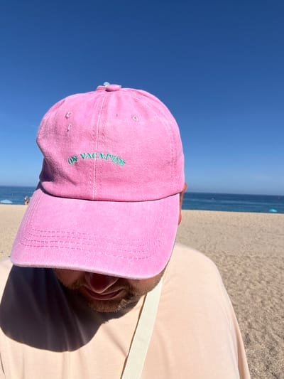 A person wearing a pink baseball cap with the words "ON VACATION" embroidered in green is lying down on a sandy beach. The cap is tilted forward, obscuring their face. The person is wearing a light-colored t-shirt. Behind them, the blue ocean meets a clear, bright blue sky, indicating it is a sunny day, likely midday given the strong shadows. The scene is set at Platja D'aro, Spain, a coastal town known for its beaches. The overall impression is one of relaxation and enjoyment of a holiday.
