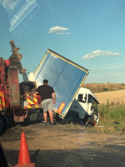 A tow truck is seen hooking up a crashed semi-truck with a blue trailer in Bozduganovo, Bulgaria. The semi-truck's cab is severely damaged, and the trailer is tilted. A man in a black t-shirt and grey shorts is standing near the rear of the tow truck, appearing to be supervising the operation. The crash site is on the side of a rural road, with a dry, harvested field in the background. A red traffic cone is visible in the foreground. The sky is blue with a few white clouds.