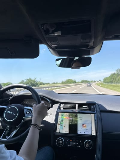 The image shows the interior of a Jaguar car, with the dashboard, steering wheel, and infotainment system visible. A person's hand is on the steering wheel. Outside the car, a highway stretches into the distance, with other vehicles visible. The infotainment screen displays navigation indicating a destination of Beograd, with a distance of 147 km and an ETA of 15:11. The dashboard temperature reads 22.0. The music track "Красиви лъжи" by Kristo is playing. The location is Paracin Municipality, Serbia.